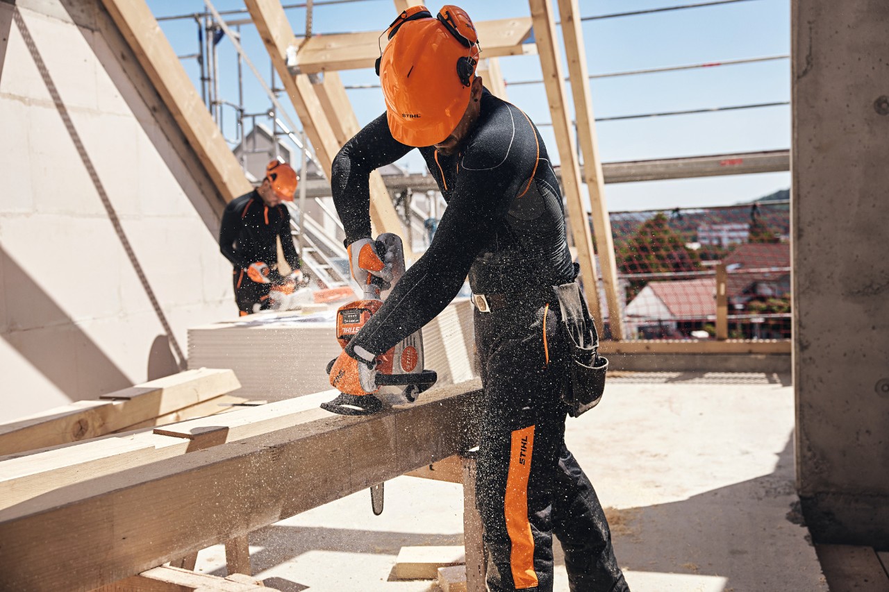 С помощта на акумулаторен моторен трион професионалист на STIHL на носещата конструкция на покрив реже дървена греда, на заден фон работи друг работник с акумулаторен моторен трион