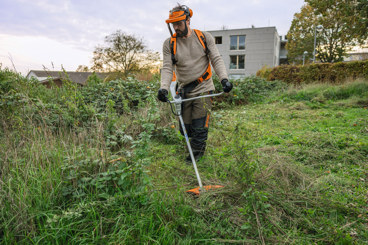 Мъж с лични предпазни средства STIHL коси зелена площ с избуяла растителност с акумулаторен храсторез STIHL FSA 200