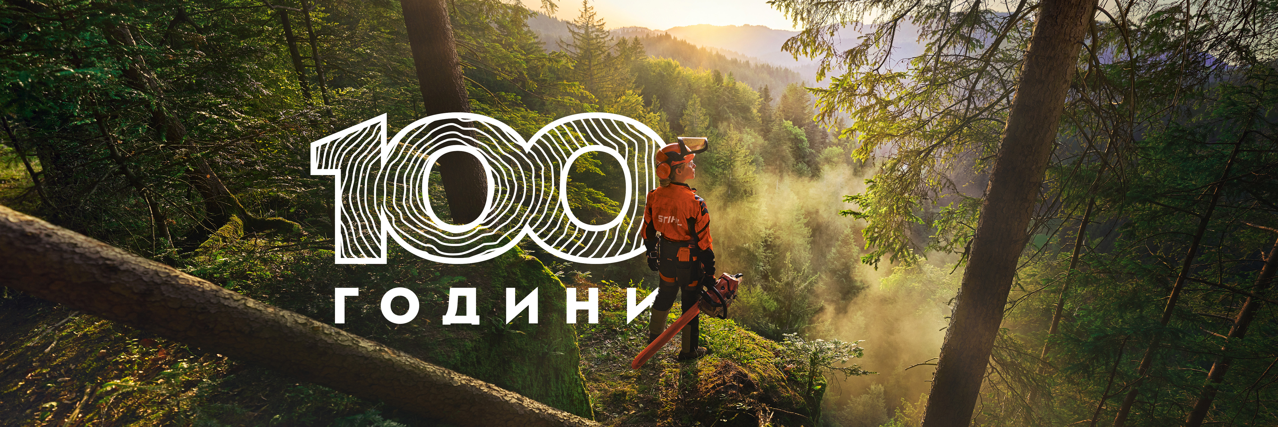 Жена стои на ръба на скала в наситено зелена гора. Тя е облечена в оранжево работно облекло STIHL, слушалки ADVANCE ProCom и държи верижен трион STIHL MS.