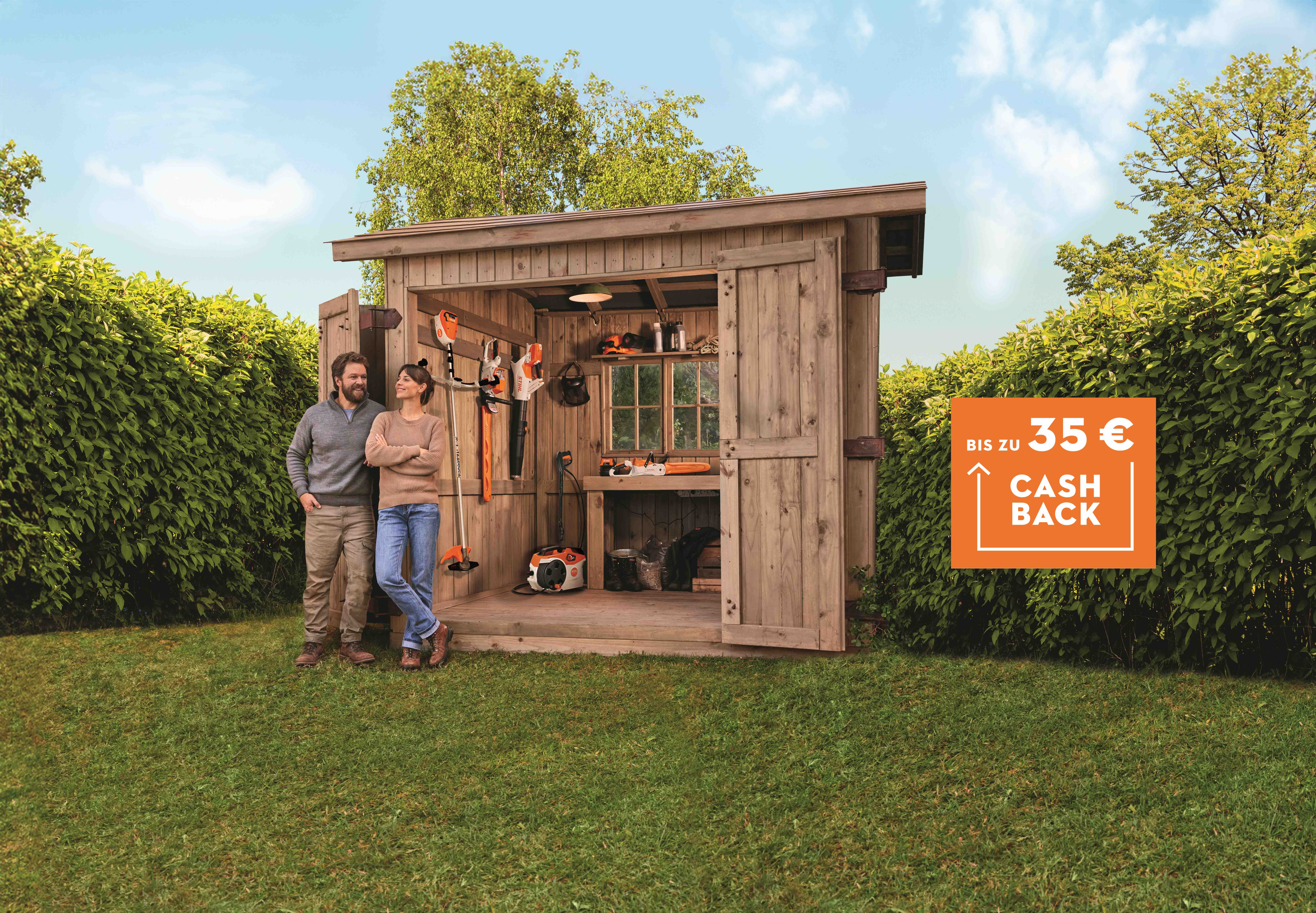 Diverse STIHL Akku Produkte aus dem Akku AK-System auf einem Rasen mit zwei Personen vor einer Holzhütte und Cashback Badge.